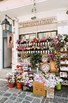 A flower shop is adorned with a vibrant display of various flowers in pots, baskets, and crates. The storefront is decorated with colorful floral arrangements and greenery. A large street lamp is mounted on the wall, and multiple signs are present with text. Several religious icons are visible in the window above the flowers.