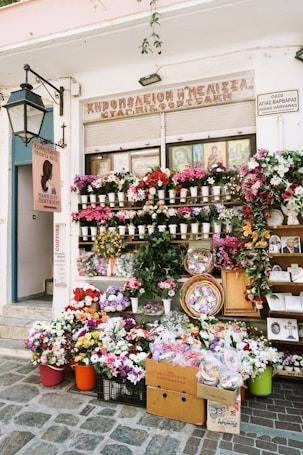 A flower shop is adorned with a vibrant display of various flowers in pots, baskets, and crates. The storefront is decorated with colorful floral arrangements and greenery. A large street lamp is mounted on the wall, and multiple signs are present with text. Several religious icons are visible in the window above the flowers.