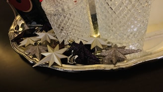 Several decorative star ornaments, both glittery and plain, rest on a reflective silver tray alongside two textured glass containers.