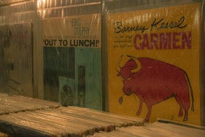 Rows of vinyl records in a dimly lit store with plastic covers. Two prominent records are visible: one by Eric Dolphy titled 'Out to Lunch!' with a subdued blue and white cover and another by Barney Kessel titled 'Carmen' featuring a bright yellow cover with a red bull.