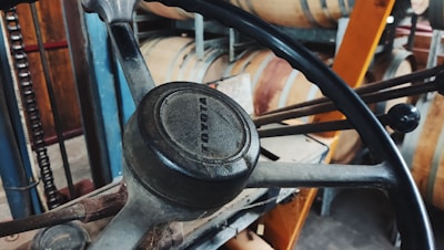 A close-up view of a steering wheel with a worn appearance, featuring the Toyota logo prominently in the center. In the background, wooden barrels and industrial equipment are visible.