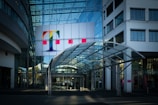 A modern office building with a prominent glass facade and a large colorful logo featuring a letter 'T' on the upper section. The entrance is covered by a transparent canopy supported by metal beams, leading into the building. The architecture displays a sleek, contemporary design with a mix of glass and concrete elements. Large windows are visible on both sides of the structure.