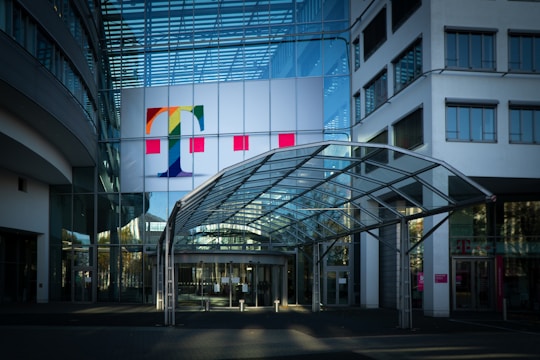 A modern office building with a prominent glass facade and a large colorful logo featuring a letter 'T' on the upper section. The entrance is covered by a transparent canopy supported by metal beams, leading into the building. The architecture displays a sleek, contemporary design with a mix of glass and concrete elements. Large windows are visible on both sides of the structure.