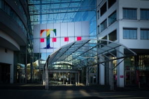A modern office building with a prominent glass facade and a large colorful logo featuring a letter 'T' on the upper section. The entrance is covered by a transparent canopy supported by metal beams, leading into the building. The architecture displays a sleek, contemporary design with a mix of glass and concrete elements. Large windows are visible on both sides of the structure.