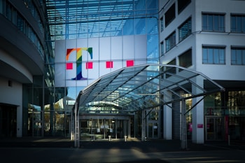 A modern office building with a prominent glass facade and a large colorful logo featuring a letter 'T' on the upper section. The entrance is covered by a transparent canopy supported by metal beams, leading into the building. The architecture displays a sleek, contemporary design with a mix of glass and concrete elements. Large windows are visible on both sides of the structure.