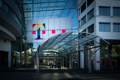 A modern office building with a prominent glass facade and a large colorful logo featuring a letter 'T' on the upper section. The entrance is covered by a transparent canopy supported by metal beams, leading into the building. The architecture displays a sleek, contemporary design with a mix of glass and concrete elements. Large windows are visible on both sides of the structure.