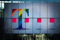 A large glass facade displays a colorful letter T and two red rectangular shapes. The letter T is decorated with a vibrant rainbow pattern, set against a light gray background.