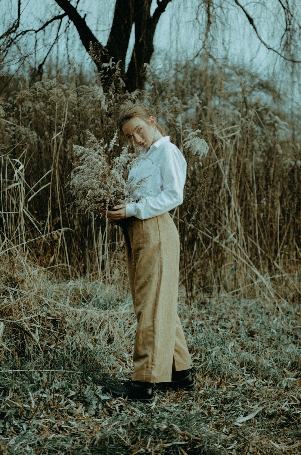 woman in white long sleeve shirt and brown pants standing on dried grass during daytime