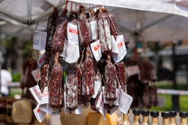 A clustered display of cured meat sausages hanging from strings, each wrapped with labels under a tent at an outdoor market. Below, various bottles with dark caps are visible, likely containing condiments.