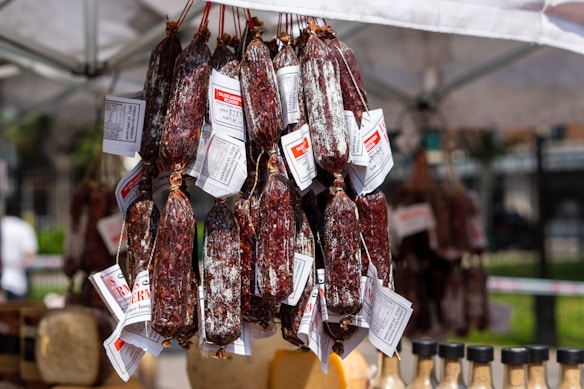 A clustered display of cured meat sausages hanging from strings, each wrapped with labels under a tent at an outdoor market. Below, various bottles with dark caps are visible, likely containing condiments.