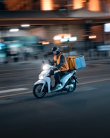 A person rides a white scooter while wearing a helmet and an orange reflective vest. The scooter has a large insulated box mounted at the back, suggesting food delivery. The background is blurred, indicating motion and a city setting with streetlights and buildings.