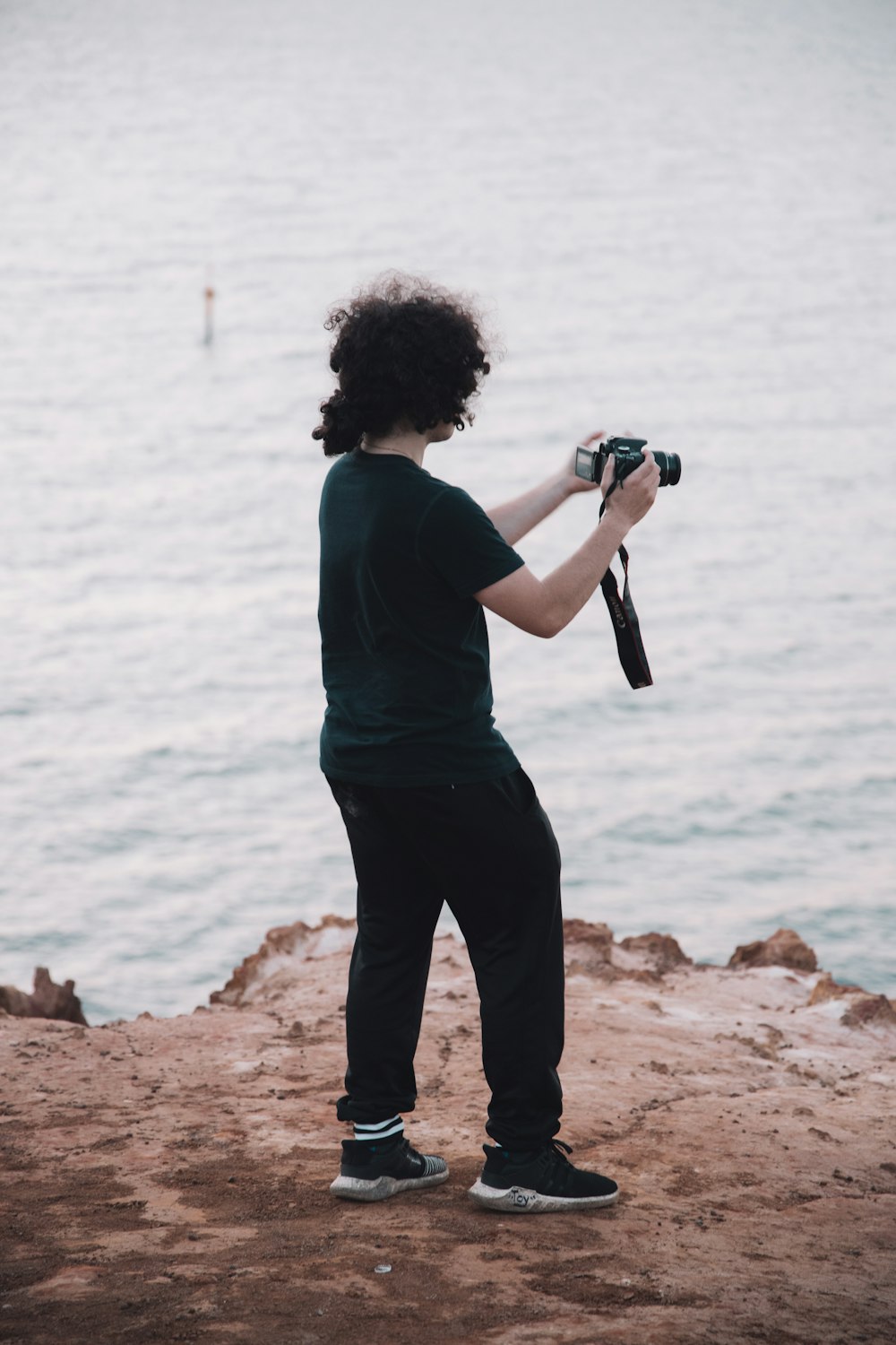 man in blue shirt and black pants holding camera standing on rock near body of water