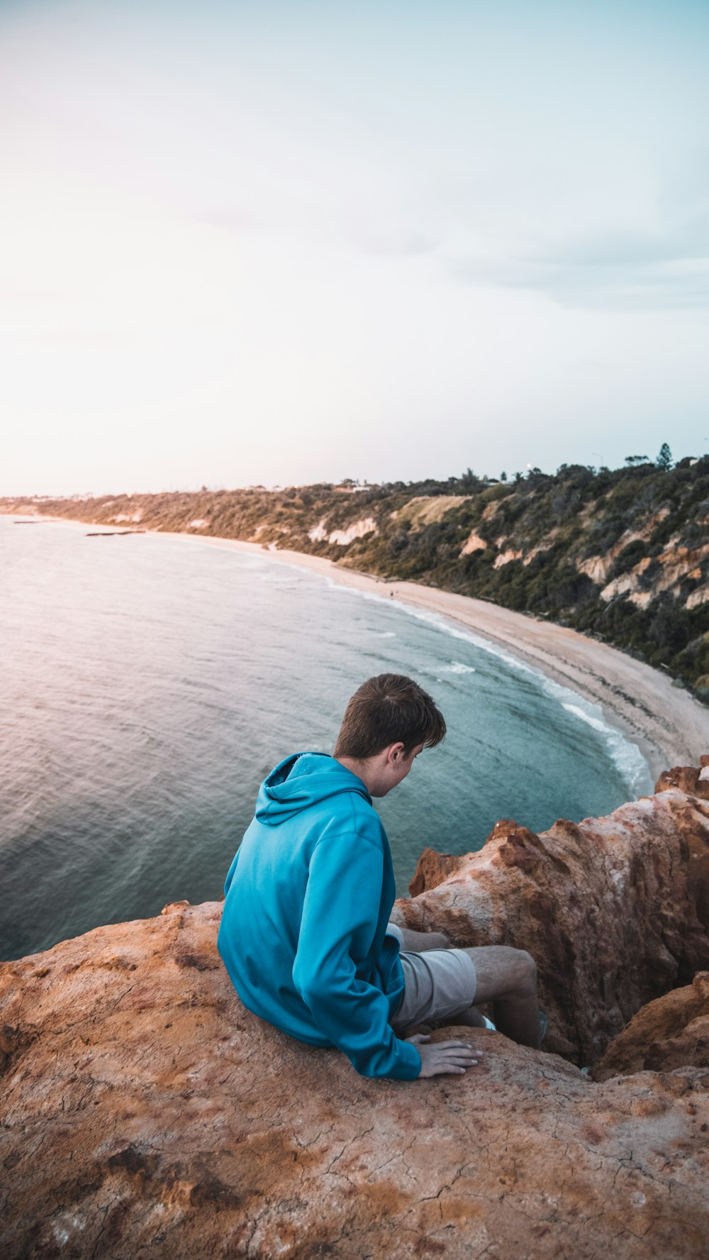 man in blue hoodie sitting on rock formation looking at sea during daytime