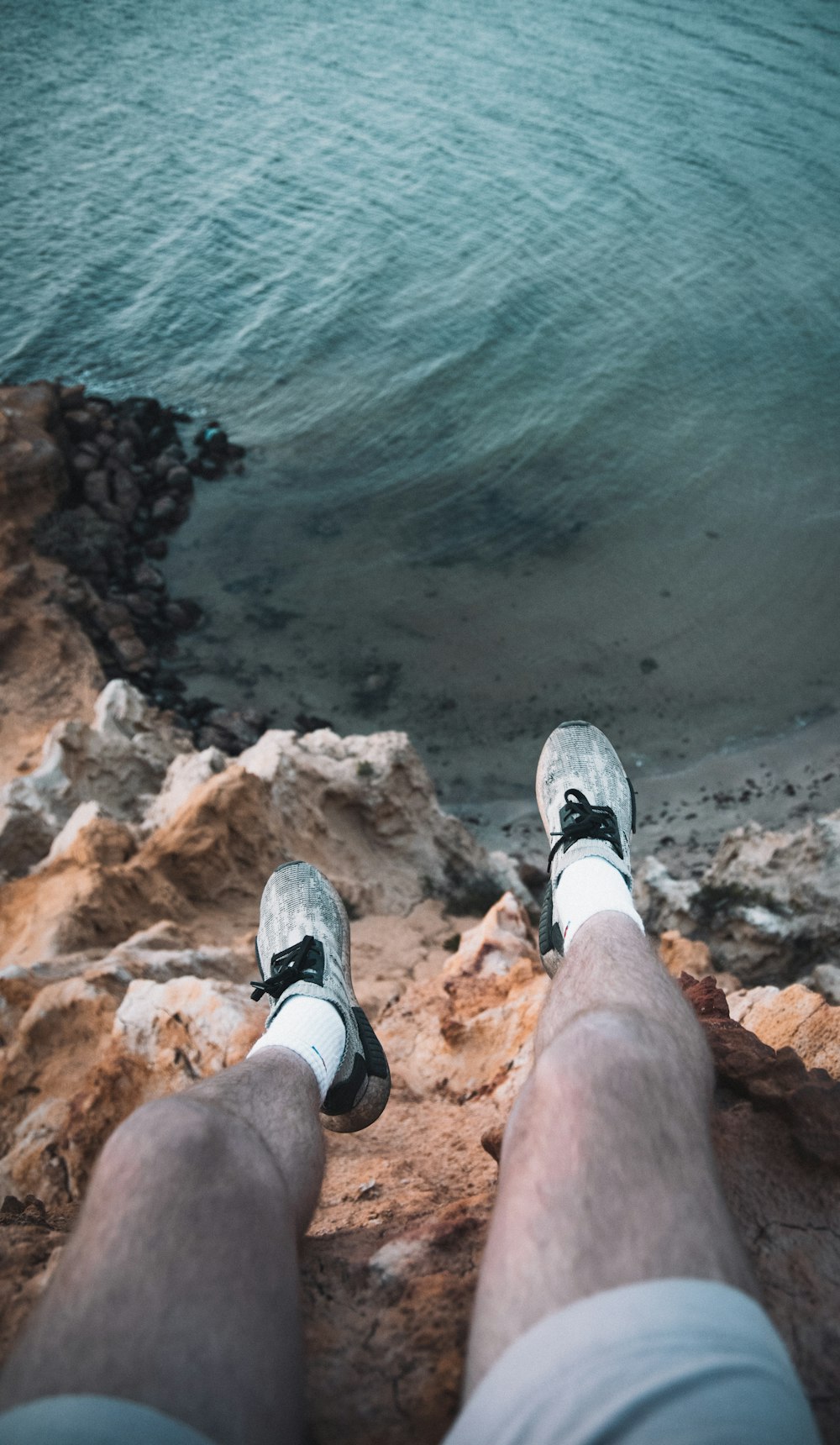 person in black and white sneakers sitting on brown rock formation near body of water during