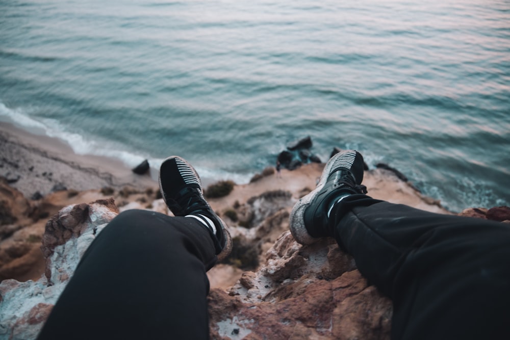 person in black pants and black and white sneakers sitting on rock near body of water