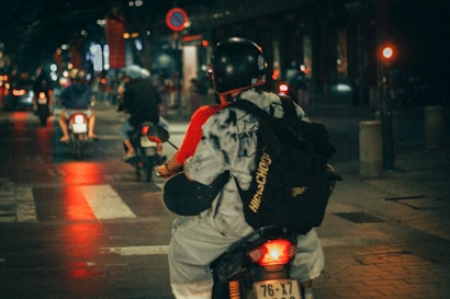 A person wearing a helmet and backpack is riding a motorbike on a city street at night. Surrounding the rider are other motorcyclists, with blurred motion and lights creating a dynamic, urban atmosphere. The street is illuminated by streetlights and the glow of red and orange reflections.