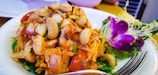 A dish featuring a colorful mix of fresh vegetables, seafood, and nuts, garnished with vibrant edible flowers and herbs on the side. A fork is placed in the foreground of the plate.