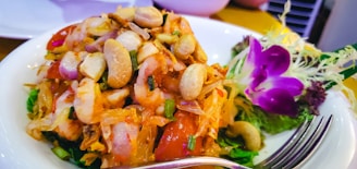A dish featuring a colorful mix of fresh vegetables, seafood, and nuts, garnished with vibrant edible flowers and herbs on the side. A fork is placed in the foreground of the plate.
