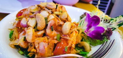 A dish featuring a colorful mix of fresh vegetables, seafood, and nuts, garnished with vibrant edible flowers and herbs on the side. A fork is placed in the foreground of the plate.