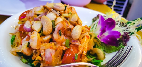 A dish featuring a colorful mix of fresh vegetables, seafood, and nuts, garnished with vibrant edible flowers and herbs on the side. A fork is placed in the foreground of the plate.