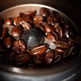Close-up shot of dark roasted coffee beans inside a metal coffee grinder. The beans are glossy and rich in color, surrounding the central blade of the grinder.