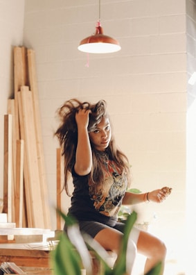 A woman with long hair is sitting on a stool in a well-lit room with a rustic decor. She is wearing a printed t-shirt and shorts. Her hand is touching her hair, and she looks directly at the camera. There are wooden planks leaning against the wall, and a pendant light hangs from the ceiling.