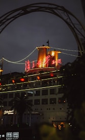 A large cruise ship with brightly illuminated signage in red, surrounded by decorative string lights. The scene is framed with an arch, and a palm tree is visible in the foreground. The atmosphere appears to be during dusk or early evening with soft ambient lighting.