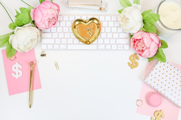 A flatlay of an organized and aesthetically pleasing workspace. Floral elements with pink and white peonies complement a gold heart-shaped trinket dish placed on a white keyboard. Additional elements include a pink notepad with a dollar sign, gold pen, paper clips, a candle, and a gift box with a white and gold design. The overall setup is bright and feminine.