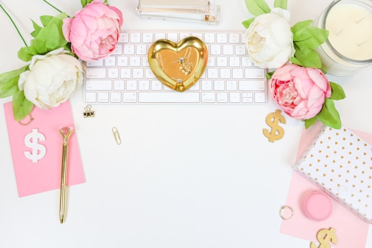A flatlay of an organized and aesthetically pleasing workspace. Floral elements with pink and white peonies complement a gold heart-shaped trinket dish placed on a white keyboard. Additional elements include a pink notepad with a dollar sign, gold pen, paper clips, a candle, and a gift box with a white and gold design. The overall setup is bright and feminine.