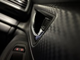 A close-up view of a car interior, focusing on a textured panel with a pattern resembling carbon fiber. The image features part of a car door with a chrome-accented handle and nearby buttons, possibly for window or lock controls. The overall look is sleek and modern.