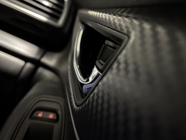 A close-up view of a car interior, focusing on a textured panel with a pattern resembling carbon fiber. The image features part of a car door with a chrome-accented handle and nearby buttons, possibly for window or lock controls. The overall look is sleek and modern.
