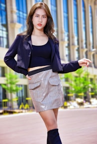 A fashionable individual stands confidently in an urban setting. The person is wearing a stylish black jacket, a black top, and a plaid skirt. Their outfit is completed with high black boots. The background features modern architecture with tall glass windows and some greenery.
