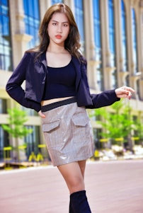 A fashionable individual stands confidently in an urban setting. The person is wearing a stylish black jacket, a black top, and a plaid skirt. Their outfit is completed with high black boots. The background features modern architecture with tall glass windows and some greenery.