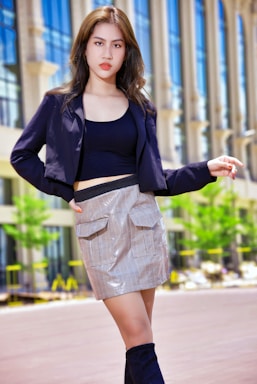 A fashionable individual stands confidently in an urban setting. The person is wearing a stylish black jacket, a black top, and a plaid skirt. Their outfit is completed with high black boots. The background features modern architecture with tall glass windows and some greenery.