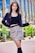 A fashionable individual stands confidently in an urban setting. The person is wearing a stylish black jacket, a black top, and a plaid skirt. Their outfit is completed with high black boots. The background features modern architecture with tall glass windows and some greenery.
