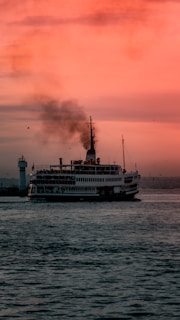 A ferryboat sailing on a calm sea during sunset, with a lighthouse visible in the background. The sky is painted in shades of orange and pink while smoke billows from the boat's chimney. Seagulls are flying around, adding to the maritime atmosphere.