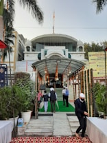 A religious building entrance is adorned with marigold garlands and greenery. Several people, some wearing head coverings, are walking towards the entrance. The building has a domed top and a prominent flag on the roof. Signage in the area indicates it is a Gurdwara, and there are palm trees and potted plants along the pathway.