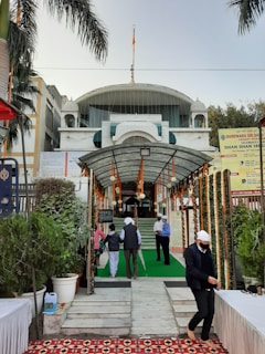 A religious building entrance is adorned with marigold garlands and greenery. Several people, some wearing head coverings, are walking towards the entrance. The building has a domed top and a prominent flag on the roof. Signage in the area indicates it is a Gurdwara, and there are palm trees and potted plants along the pathway.