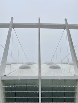 A modern architectural structure with a series of large, white, tent-like canopies supported by steel cables and beams. The canopies have dome-shaped tops and the structure appears minimalistic and geometric.