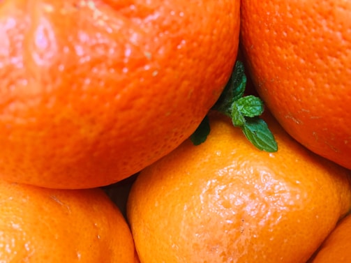 A close-up view of several ripe oranges with a shiny, textured peel. The bright orange color is contrasted by small green leaves peeking through.