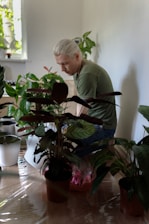 A person with light-colored hair is focused on a task, surrounded by several potted plants indoors. They are wearing a green shirt and blue jeans, kneeling on a hardwood floor. Sunlight filters in from a window, casting light on the plants and creating a calm atmosphere.