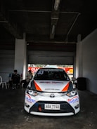 A white race car with various sponsor logos and the number 53 prominently displayed is parked in a dimly lit garage or pit area. The car features orange and blue accents and a visible Toyota emblem on the front. Two people are sitting at a table to the side, and tires are stacked against the wall.