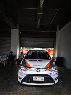 A white race car with various sponsor logos and the number 53 prominently displayed is parked in a dimly lit garage or pit area. The car features orange and blue accents and a visible Toyota emblem on the front. Two people are sitting at a table to the side, and tires are stacked against the wall.