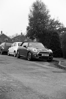 Several cars are parked along the side of a residential street. In the foreground, a dark-colored Mini Cooper with the license plate GX16 YHT is prominently visible, while trees and houses provide a suburban backdrop.