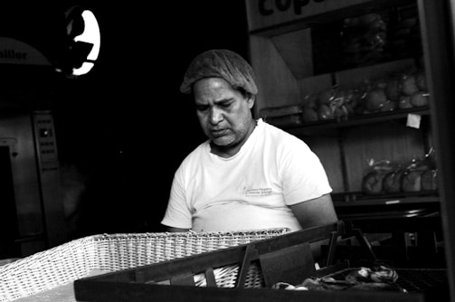 A man wearing a hairnet and a white t-shirt stands behind a basket in a bakery. The shelves in the background are lined with loaves of bread wrapped in plastic.