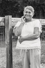 An elderly woman is leaning against a wooden fence, smiling gently. She is wearing a short-sleeved blouse with lace trim and light pants. Her hand rests on her face while the other arm is crossed. The setting appears to be outdoors, suggesting a rural or garden environment.