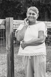 An elderly woman is leaning against a wooden fence, smiling gently. She is wearing a short-sleeved blouse with lace trim and light pants. Her hand rests on her face while the other arm is crossed. The setting appears to be outdoors, suggesting a rural or garden environment.