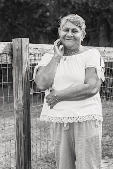 An elderly woman is leaning against a wooden fence, smiling gently. She is wearing a short-sleeved blouse with lace trim and light pants. Her hand rests on her face while the other arm is crossed. The setting appears to be outdoors, suggesting a rural or garden environment.