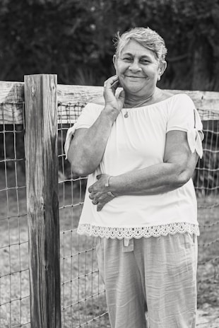 An elderly woman is leaning against a wooden fence, smiling gently. She is wearing a short-sleeved blouse with lace trim and light pants. Her hand rests on her face while the other arm is crossed. The setting appears to be outdoors, suggesting a rural or garden environment.
