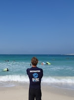 A surf instructor stands on the sandy beach with their back to the camera, observing a group of people in the ocean. Some individuals are using bodyboards on the waves. The sky is clear and blue, while the sea exhibits various shades of blue and turquoise.